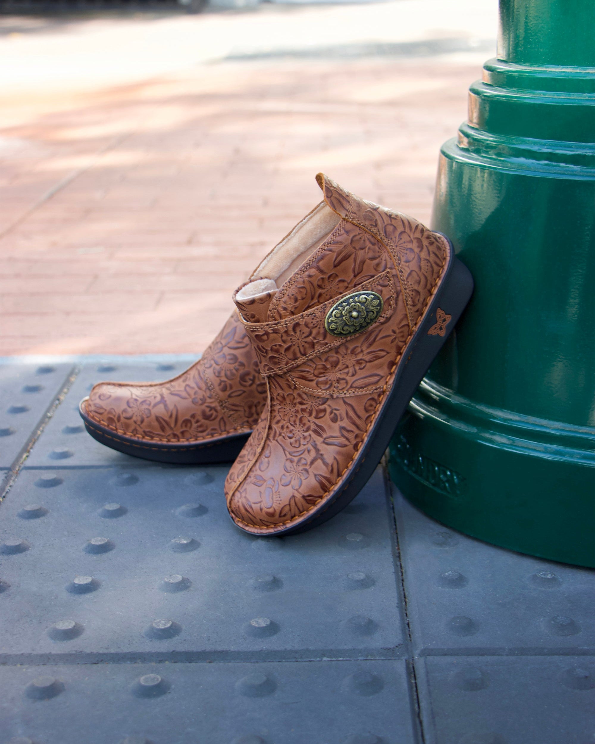 Brown leather boot with decorative emblem leaning against a green metal post on a tiled surface.