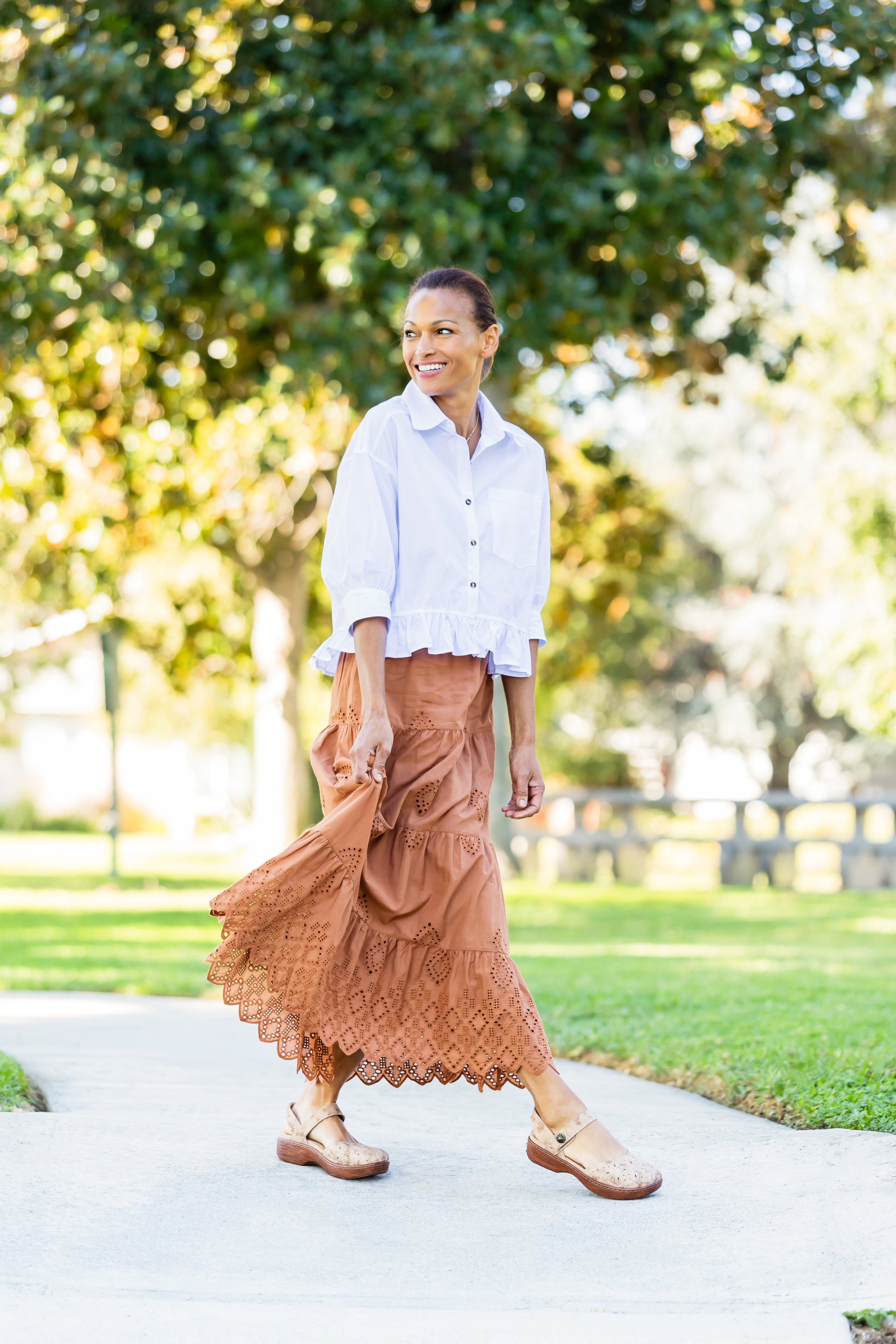 A woman walking on a paved path in a park, wearing a white button-up shirt, a rust-brown tiered maxi skirt, and brown leather shoes.