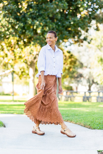 A woman walking on a paved path in a park, wearing a white button-up shirt, a rust-brown tiered maxi skirt, and brown leather shoes.