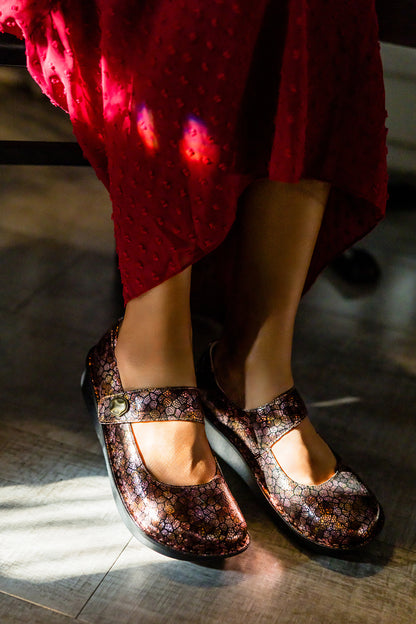 A close-up of a person wearing dark brown Mary Jane shoes with a purple and gold floral pattern, paired with a red skirt, standing on a wooden floor.