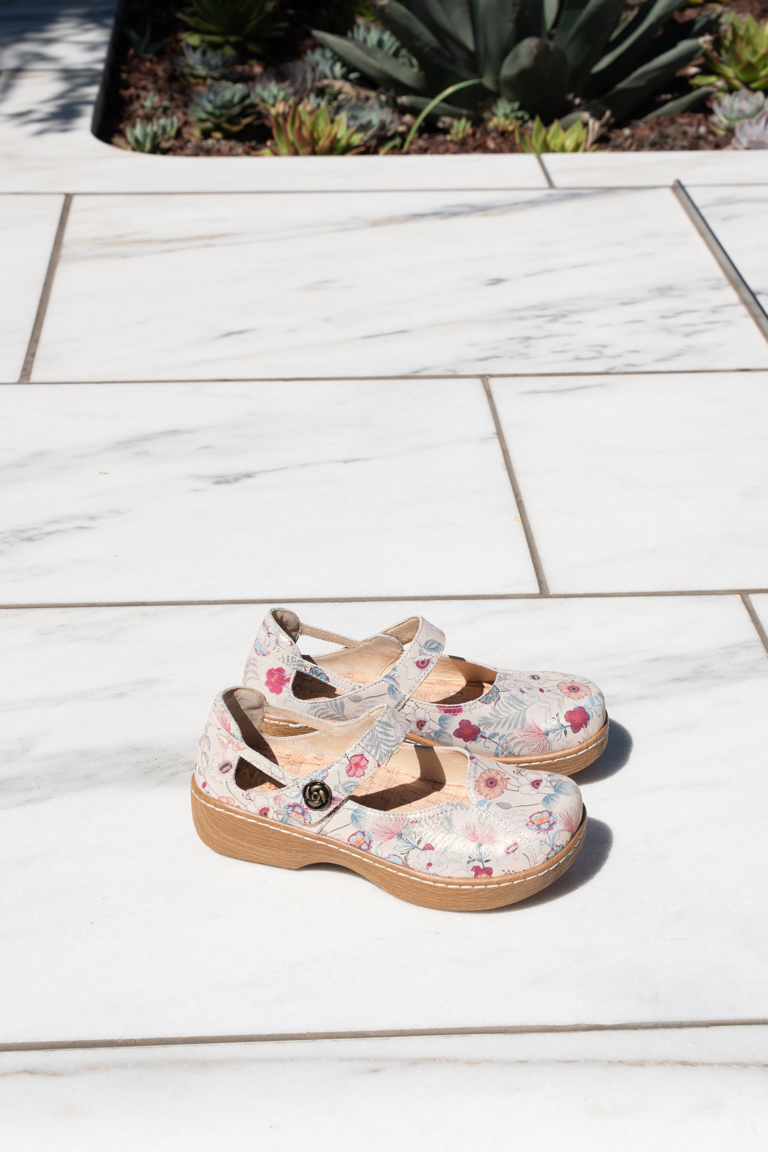 A pair of Mary Jane clogs with a light beige floral print and brown straps, resting on a white marble tile patio.
